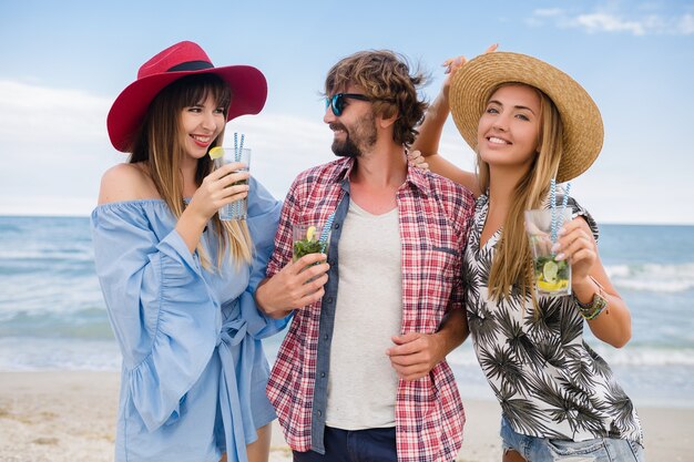 Young hipster company of friends on vacation at beach, drinking mojito cocktail, happy positive, summer style, smiling happy, two women and man having fun together, talking, flirt, romance, three