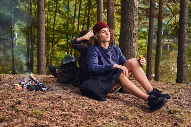 Free photo young hipster couple hiking in the forest and sitting back to back while warming near a campfire at camp. travel, tourism, and hike concept.