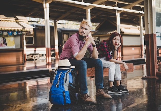 Young hipster couple sitting on wooden bench at train station. Couple sitting waiting for the train at platform.