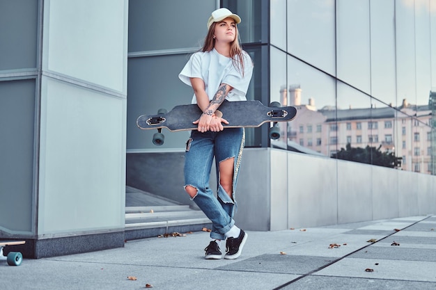 Free Photo young hipster girl in cap dressed in white shirt and ripped jeans holds skateboard while posing near skyscraper.