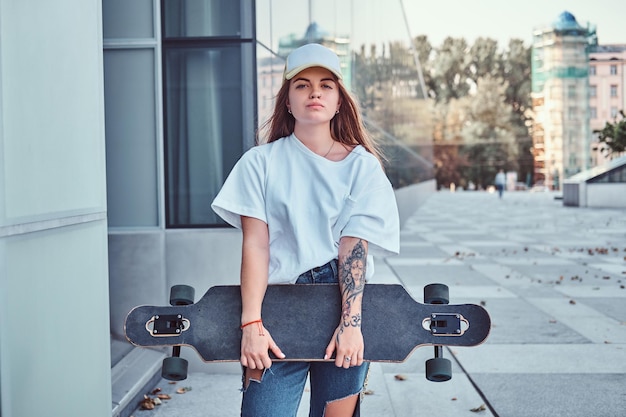 Free photo young hipster girl in cap dressed in white shirt and ripped jeans holds skateboard while posing near skyscraper.