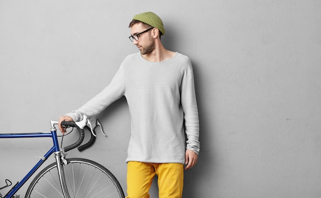 Free photo young hipster guy in loose sweater and glasses choosing bicycle for himself, wanting to exercise everyday and to have long trips on fresh air, looking at modern bicycle, keeping hand on handle bar