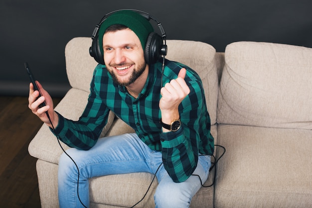 Free Photo young hipster handsome bearded man sitting on a couch at home, listening to music on headphones