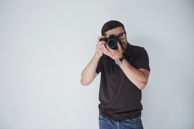 Free Photo a young hipster man in eyepieces holds a dslr camera in hands standing against a white wall