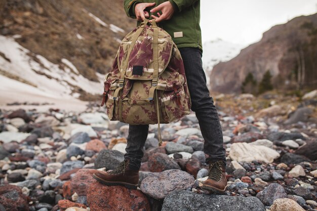 Young hipster man, hiking by the river, wild nature, winter vacation, holding backpack in hands, close up details