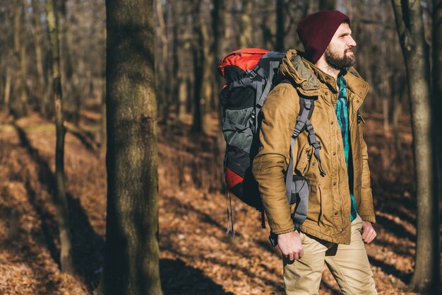 Young hipster man traveling with backpack in autumn forest wearing warm jacket and hat, active tourist, exploring nature in cold season