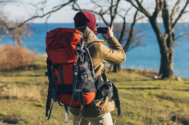 Young hipster man traveling with backpack wearing warm jacket and hat, active tourist, taking pictures on mobile phone, exploring nature in cold season