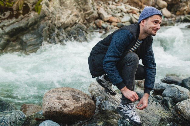 Young hipster man walking on a rock at river in winter forest, tying shoelaces