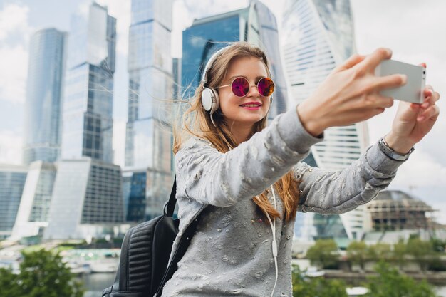 Young hipster woman having fun in street listening to music on headphones, wearing pink sunglasses, spring summer urban style, taking selfie pisture on smartphone