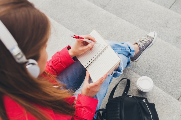 Free photo young hipster woman in pink coat, jeans sitting in street with backpack and coffee listening to music on headphones