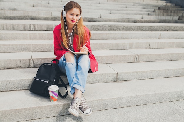 Free Photo young hipster woman in pink coat, jeans sitting in street with backpack and coffee listening to music on headphones