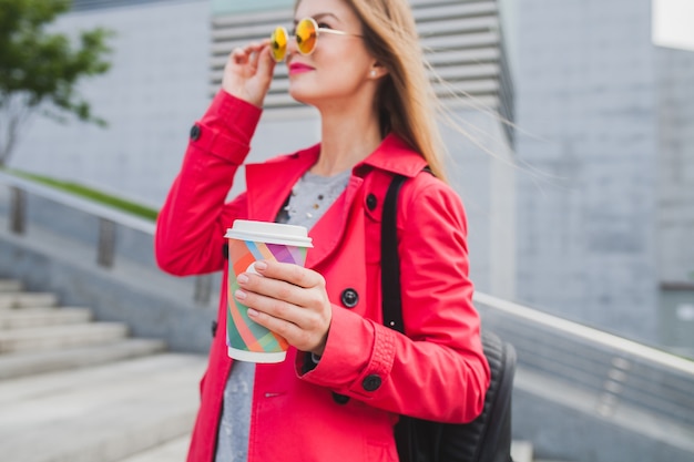 Young hipster woman in pink coat, jeans in street with backpack and coffee listening to music on headphones