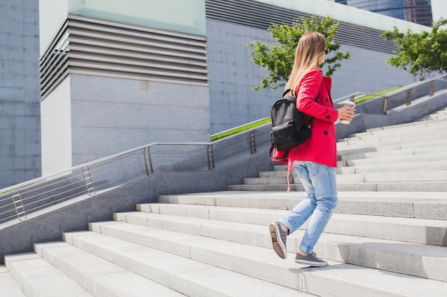 Free photo young hipster woman in pink coat, jeans walking on stairs in street with backpack and coffee listening to music on headphones, wearing sunglasses