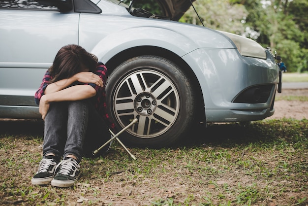 Young hipster woman waiting for roadside assistance after her car breaks down at the side of the road sitting against her car.