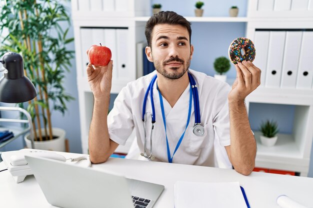 Young hispanic dietitian man holding doughnut and apple smiling looking to the side and staring away thinking.