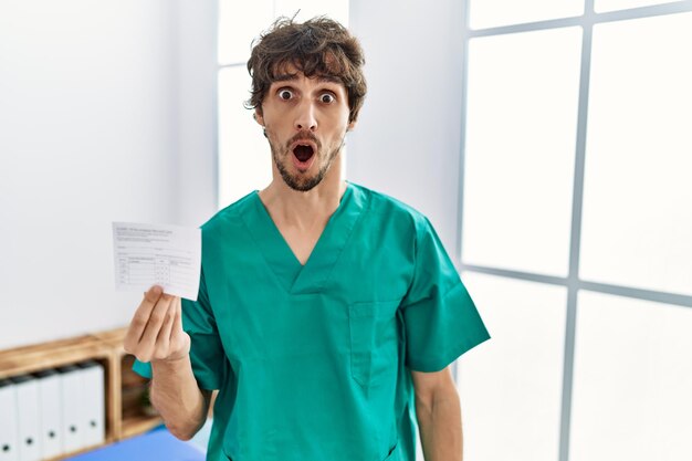 Young hispanic doctor man holding covid record card in shock face looking skeptical and sarcastic surprised with open mouth