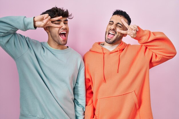 Young hispanic gay couple standing over pink background doing peace symbol with fingers over face, smiling cheerful showing victory