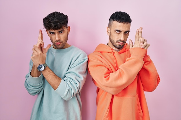 Free photo young hispanic gay couple standing over pink background holding symbolic gun with hand gesture playing killing shooting weapons angry face