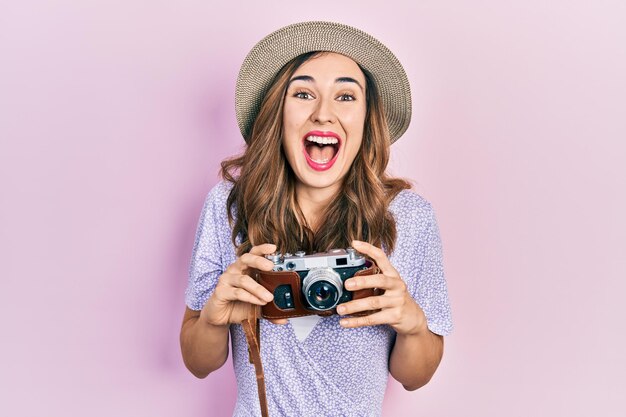 Young hispanic girl wearing summer hat holding vintage camera celebrating crazy and amazed for success with open eyes screaming excited