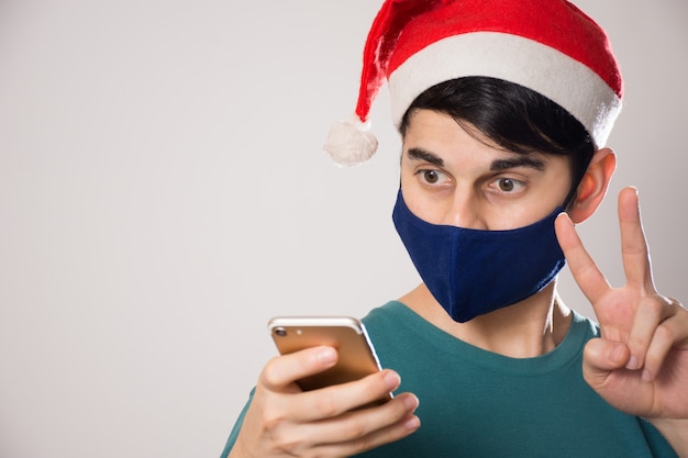 Young Hispanic male with a face mask and a Santa hat looking to his phone and doing a peace gesture