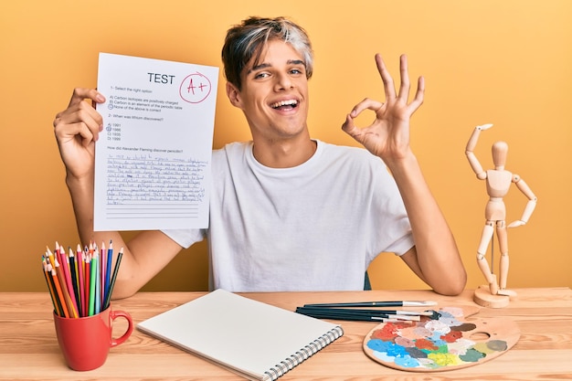 Free photo young hispanic man artist holding passed exam sitting on the table doing ok sign with fingers smiling friendly gesturing excellent symbol