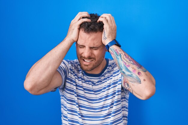 Young hispanic man standing over blue background suffering from headache desperate and stressed because pain and migraine. hands on head.