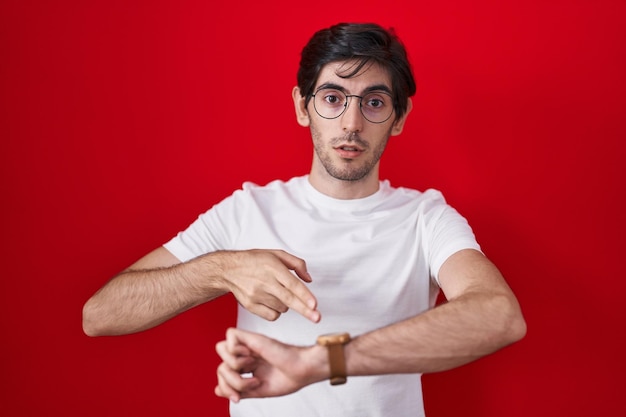 Free photo young hispanic man standing over red background in hurry pointing to watch time, impatience, upset and angry for deadline delay