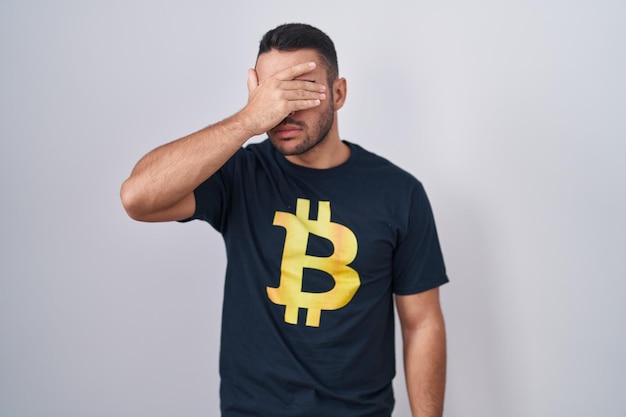 Young hispanic man wearing bitcoin t shirt covering eyes with hand looking serious and sad sightless hiding and rejection concept