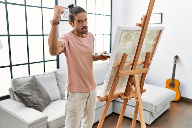 Free photo young hispanic man with beard painting on canvas at home angry and mad raising fist frustrated and furious while shouting with anger. rage and aggressive concept.