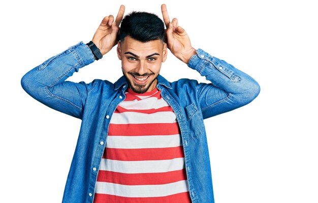 Young hispanic man with beard wearing casual denim jacket posing funny and crazy with fingers on head as bunny ears, smiling cheerful