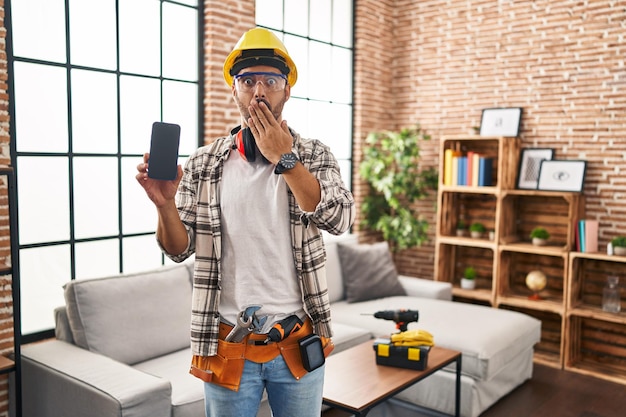 Young hispanic man with beard working at home renovation holding smartphone covering mouth with hand shocked and afraid for mistake surprised expression