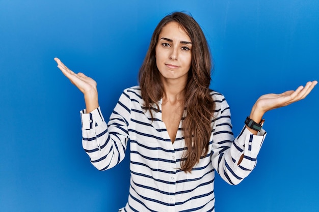 Young hispanic woman standing over blue isolated background clueless and confused expression with arms and hands raised doubt concept