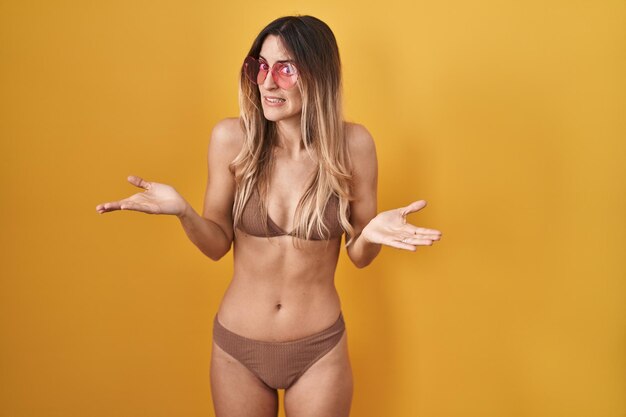 Young hispanic woman wearing bikini over yellow background clueless and confused with open arms, no idea concept.