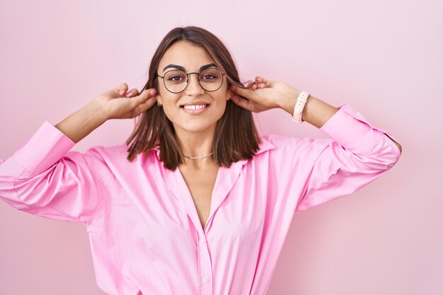 Young hispanic woman wearing glasses standing over pink background smiling pulling ears with fingers, funny gesture. audition problem