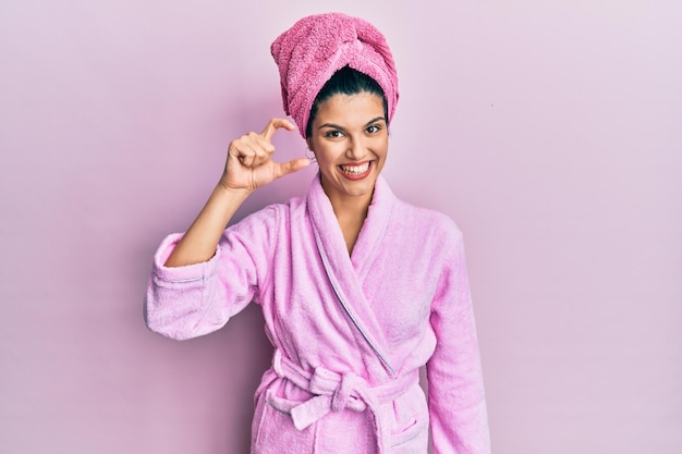 Free photo young hispanic woman wearing shower towel cap and bathrobe smiling and confident gesturing with hand doing small size sign with fingers looking and the camera measure concept