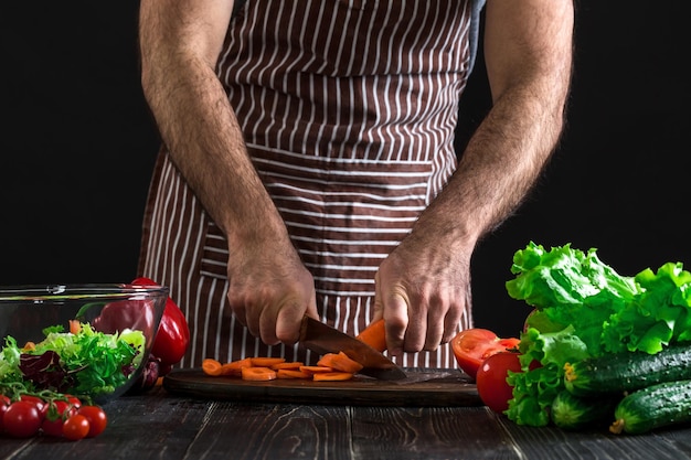 Free Photo young home cook man in apron slicing carrot with kitchen knife. men's hands cut the carrot to make a salad on black background. healthy food concept