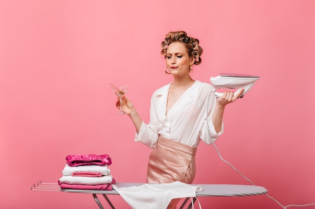 Young housewife in elegant outfit posing with iron and martini on pink wall