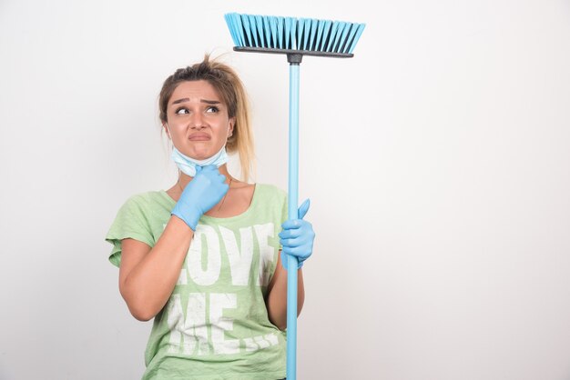 Young housewife with facemask holding broom while looking up confused.