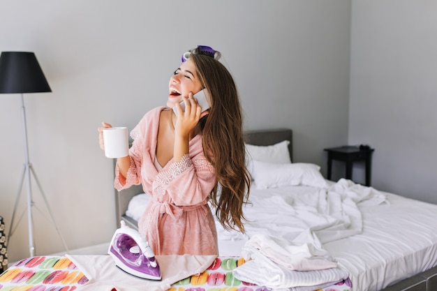 Young housewife with long hair  in pink bathrobe with curler on head at home at ironing clothes. She  speaking on phone, laughing with closed eyes.