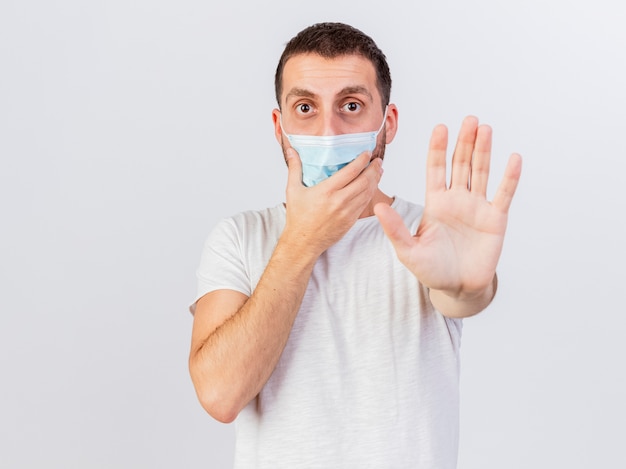 Young ill man wearing scarf and medical mask wrapped in plaid showing stop gesture isolated on white