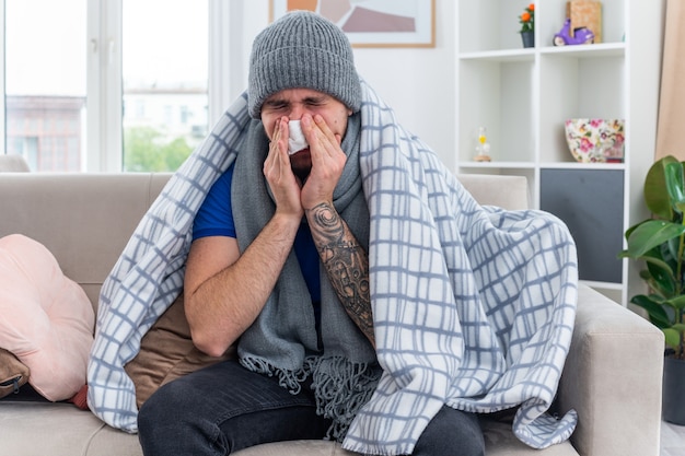 young ill man wearing scarf and winter hat wrapped in blanket sitting on sofa in living room wiping nose with napkin with closed eyes