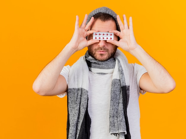 Free photo young ill man wearing winter hat and scarf covered eyes with pills isolated on yellow background