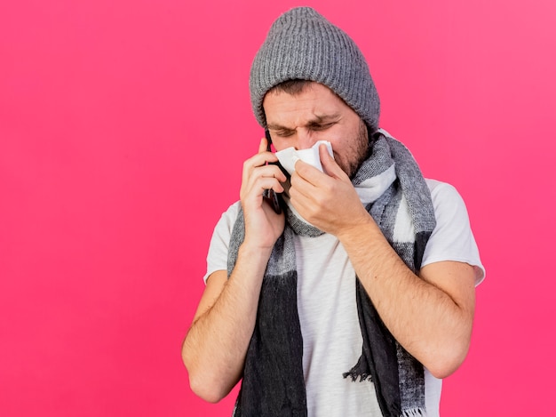 Free photo young ill man wearing winter hat with scarf speaks on phone and wiping nose with napkin isolated on pink