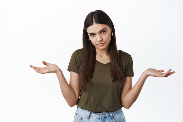 Young indifferent woman look unbothered at front, spread empty hands sideways, shrugging shoulders, standing over white wall