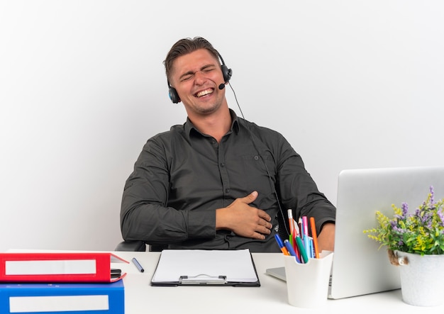 Free photo young joyful blonde office worker man on headphones sits at desk with office tools using laptop holds belly with closed eyes