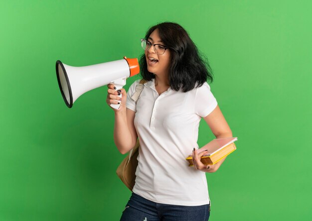 Young joyful pretty caucasian schoolgirl wearing glasses and back bag shouts through loud speaker holding books on green  with copy space