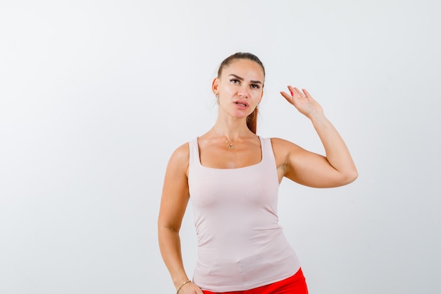 Young lady in beige tank top poising with raised hand and looking careful