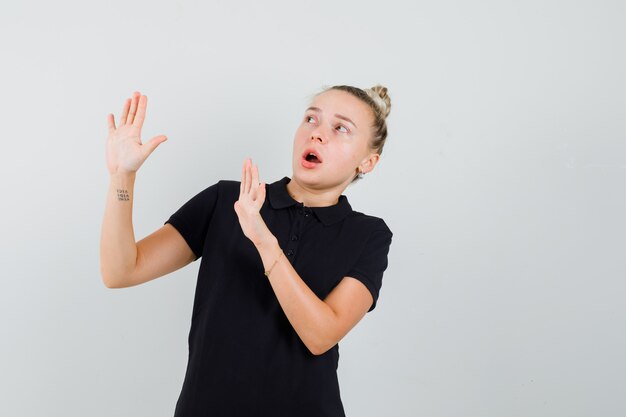 Young lady in black t-shirt raising hands to defend herself and looking scared