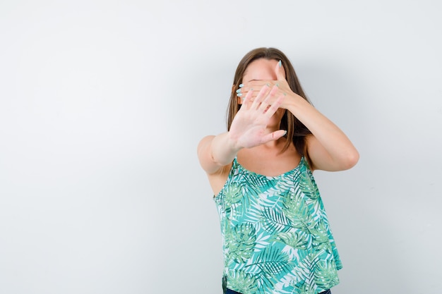 Young lady in blouse covering eyes with hand while showing stop gesture and looking serious , front view.