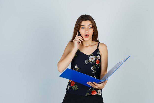 Young lady in blouse holding folder, talking on mobile phone and looking shocked , front view.